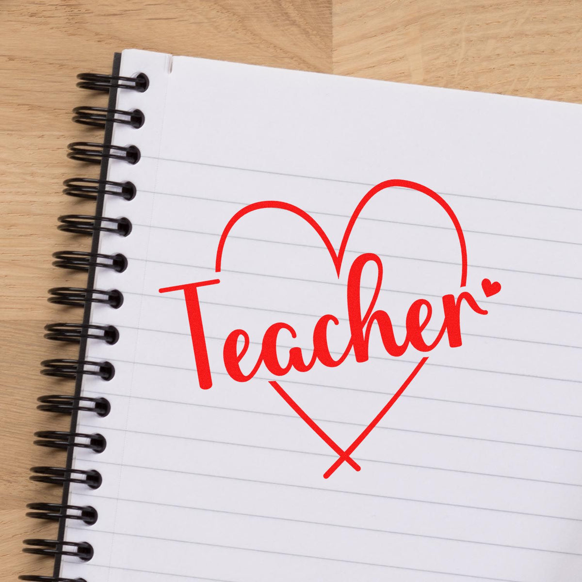 Red teacher stamp seal in a heart on a white lined spiral notebook with black binding on a wood table.