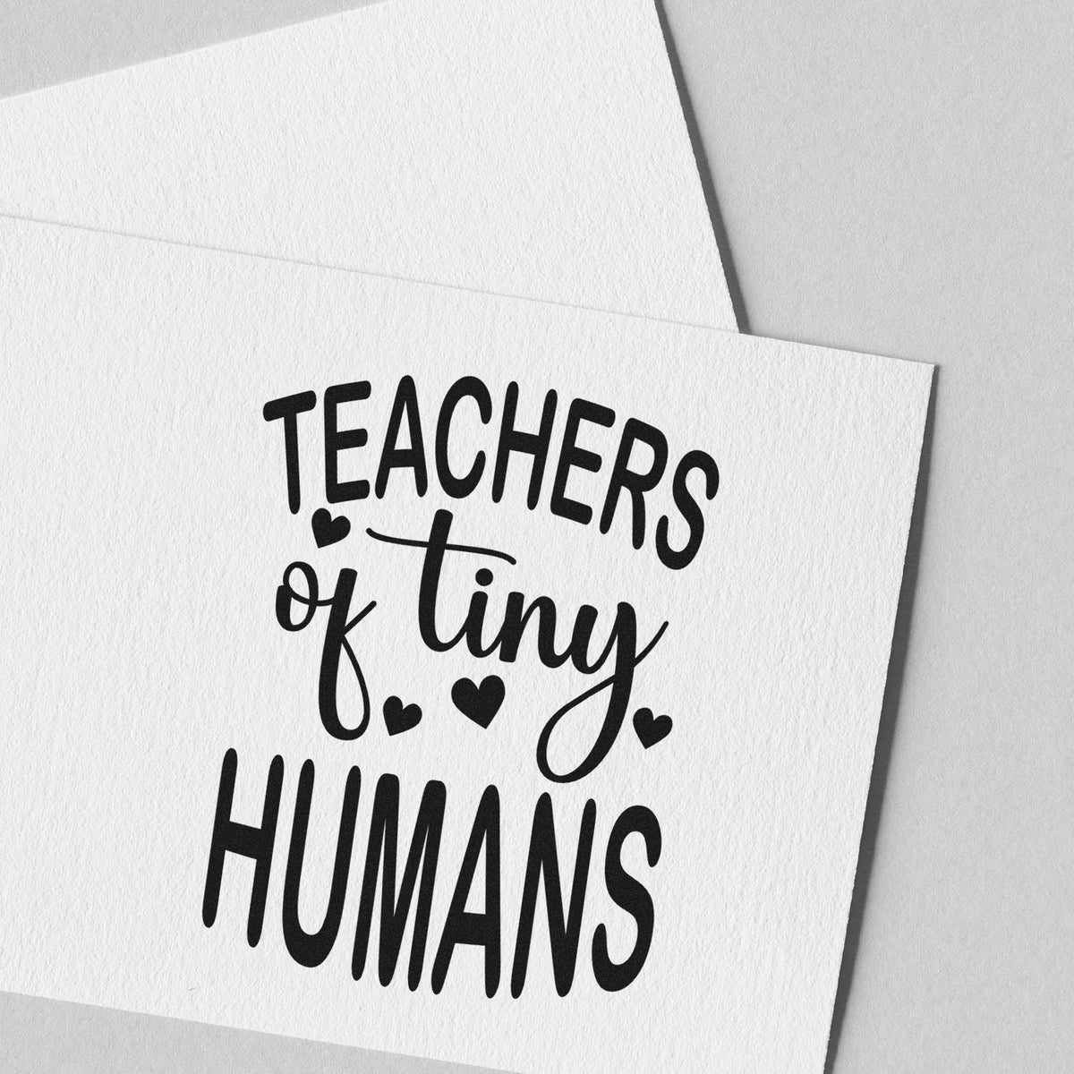 Black teacher stamp and seal on white textured paper reading Teachers of tiny humans with hearts.