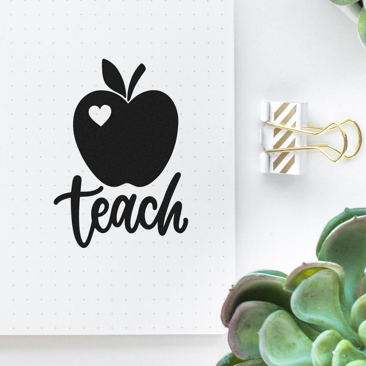 Black apple teach stamp and seal on white dotted notebook, with gold-striped binder clip and green succulent