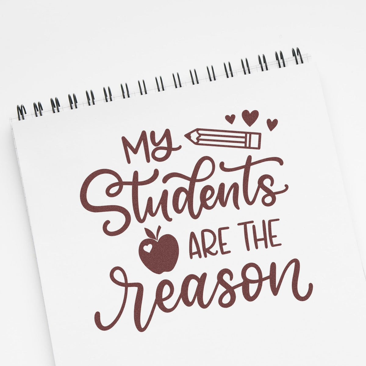 Maroon teacher stamp and seal design on spiral notebook reading My Students Are the Reason, with pencil, hearts, and apple.