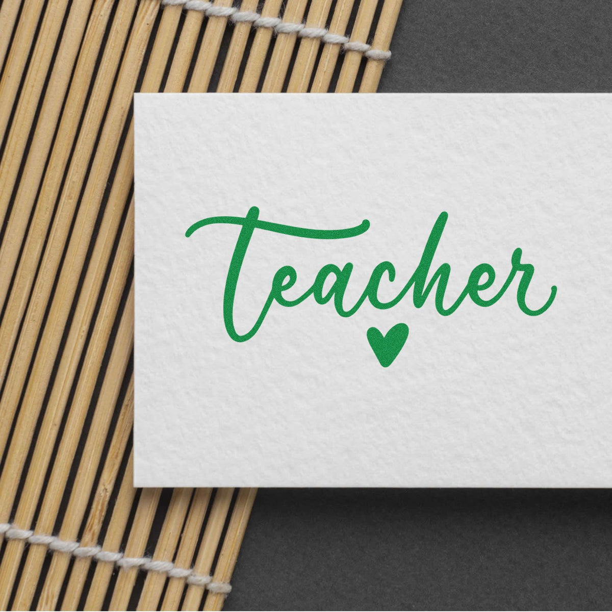 Green Teacher stamp-and-seal design with heart on white card, set on bamboo mat over a dark background.