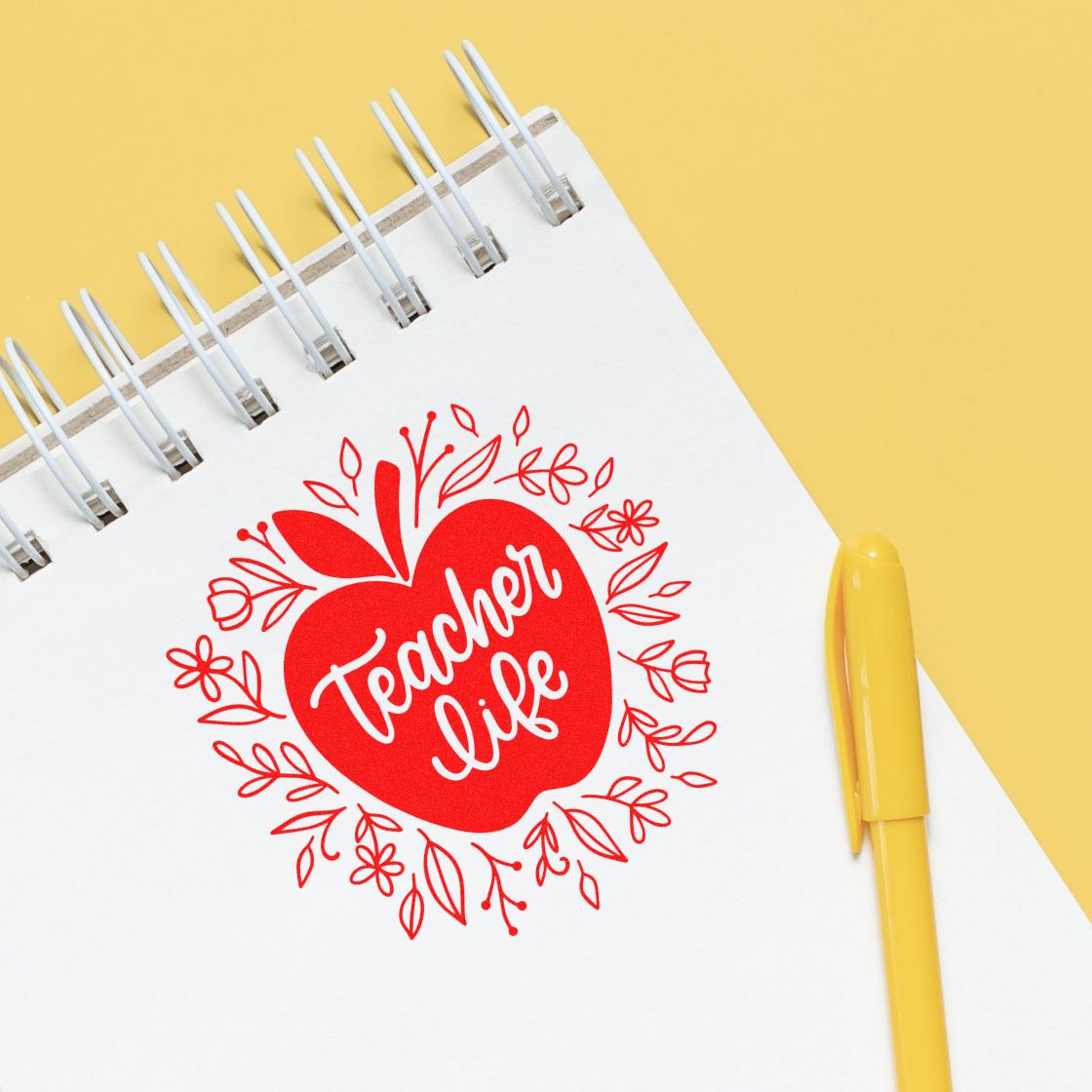 Red apple-shaped teacher life stamp and seal on a white spiral notebook with a yellow pen on a yellow background.