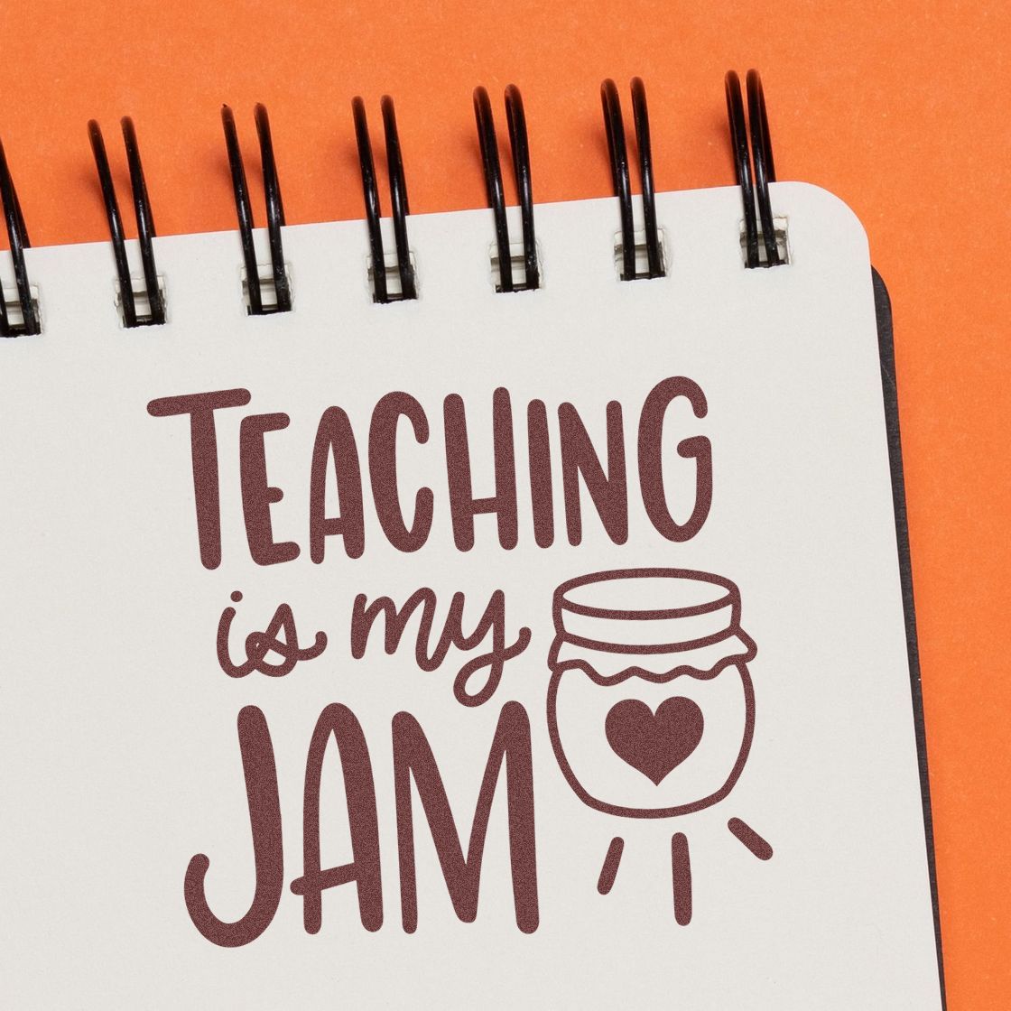 Maroon Teaching is My Jam stamp with jar-heart seal on a white spiral notebook over an orange background.