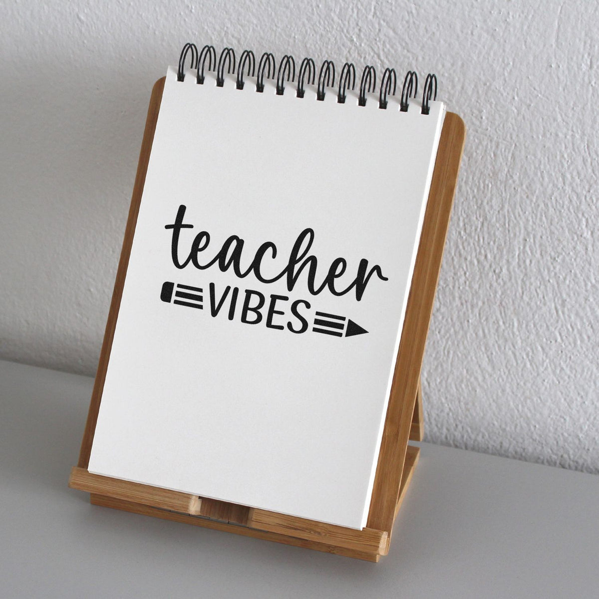 Black teacher vibes stamp impression on white spiral notepad, displayed on a wooden stand against a light gray wall.