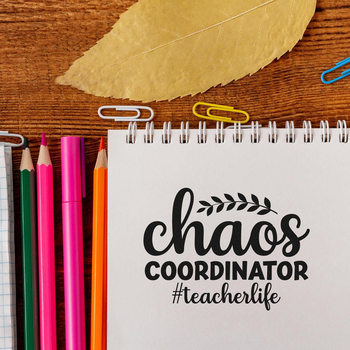 Teacher stamp/seal reading chaos coordinator #teacherlife on white spiral notebook, with colorful pencils and paper clips.