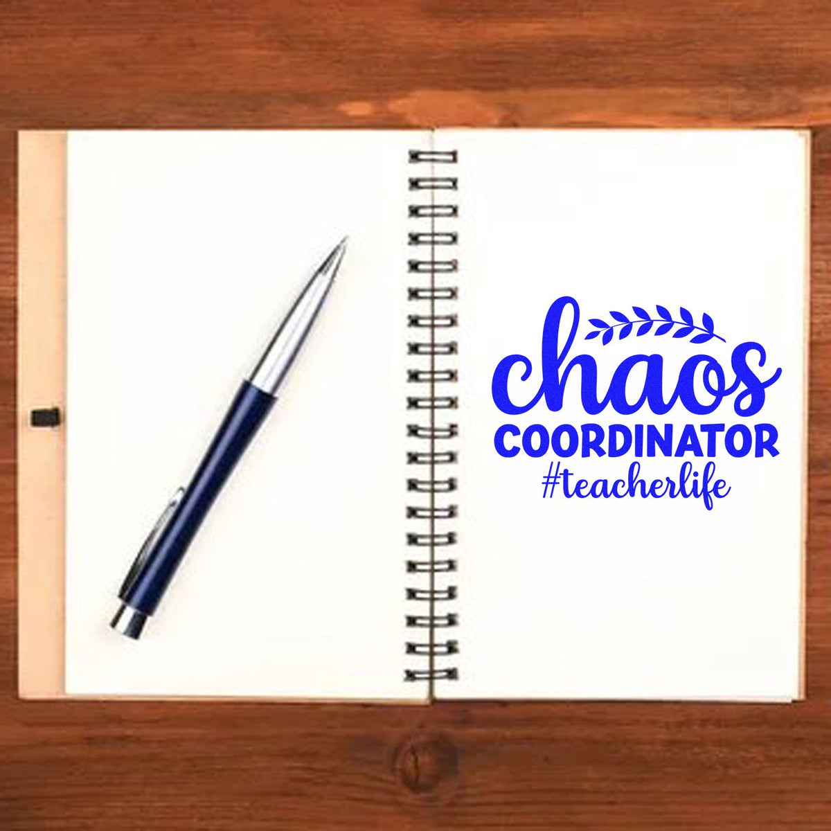 Blue chaos coordinator #teacherlife stamp/seal beside a navy pen on an open spiral notebook on a wooden desk.