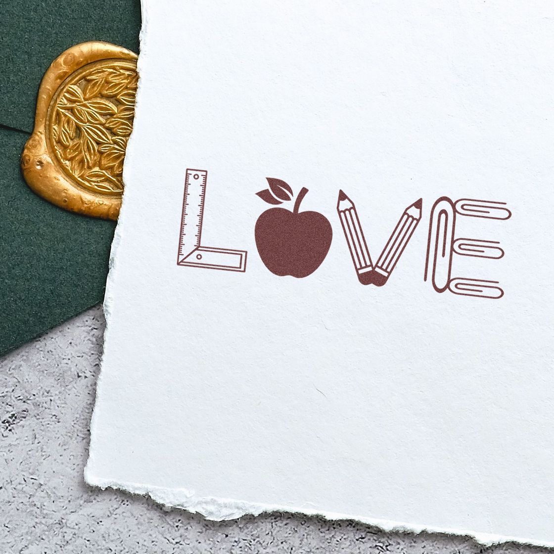 Brown LOVE stamp with ruler L, apple, pencil V and paperclip E on white paper beside a gold wax seal.