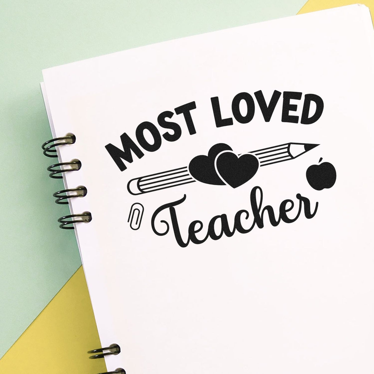 Black Most Loved Teacher stamp seal with pencil, hearts, apple, and paperclip on a white spiral notebook over pastel green and yellow