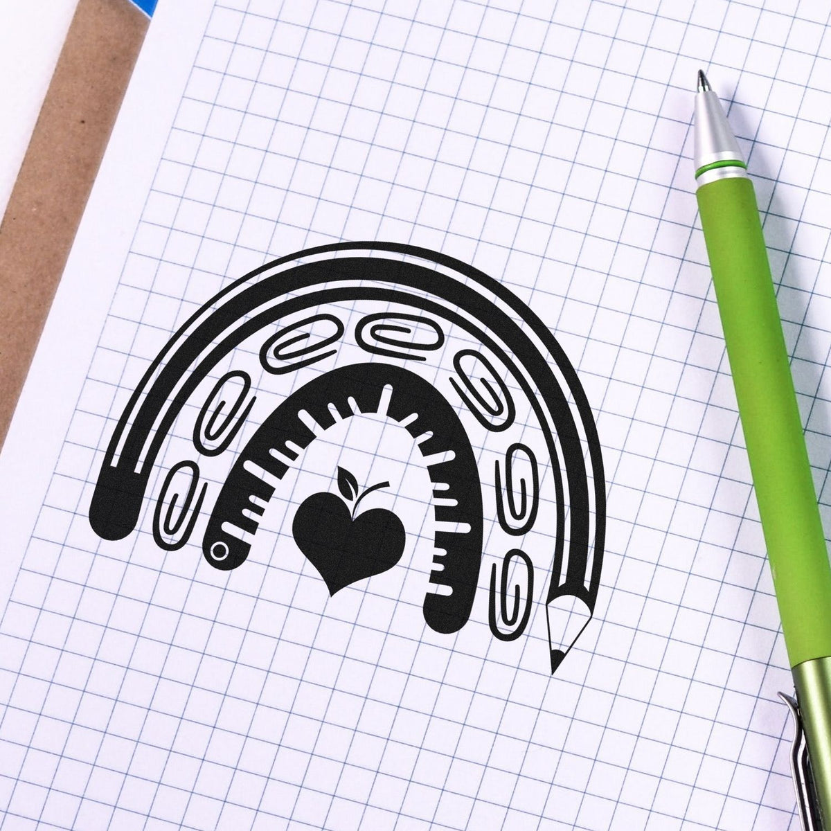 Black rainbow stamp seal with paperclips and apple icon on graph paper beside a green pen.