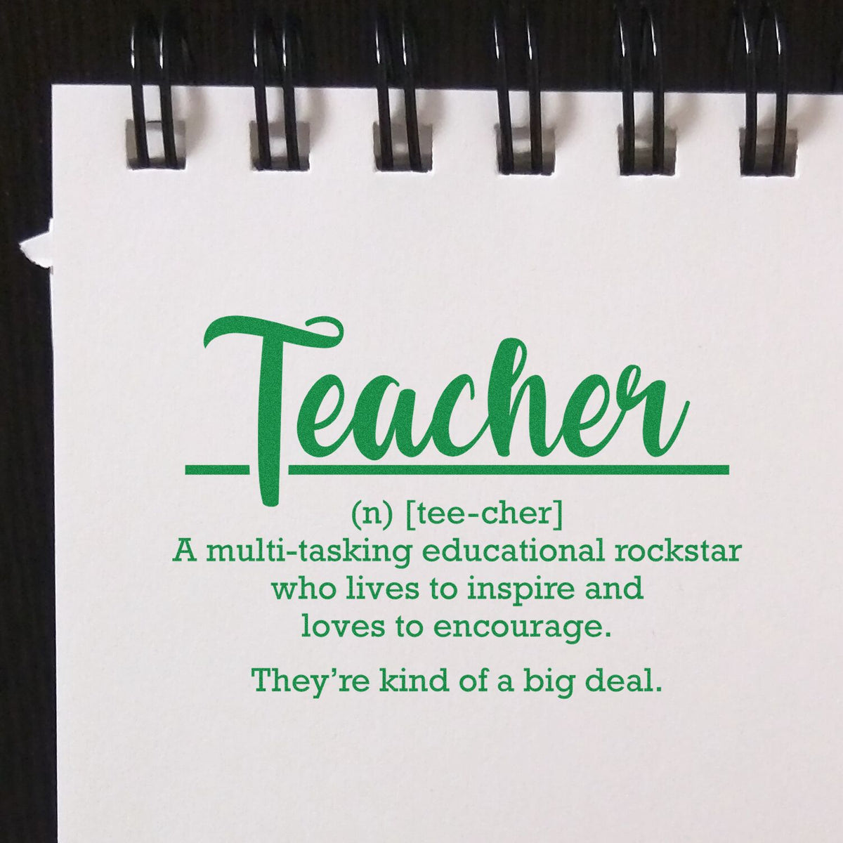 Green teacher stamp seal on white spiral notebook page, with black binding and definition text.