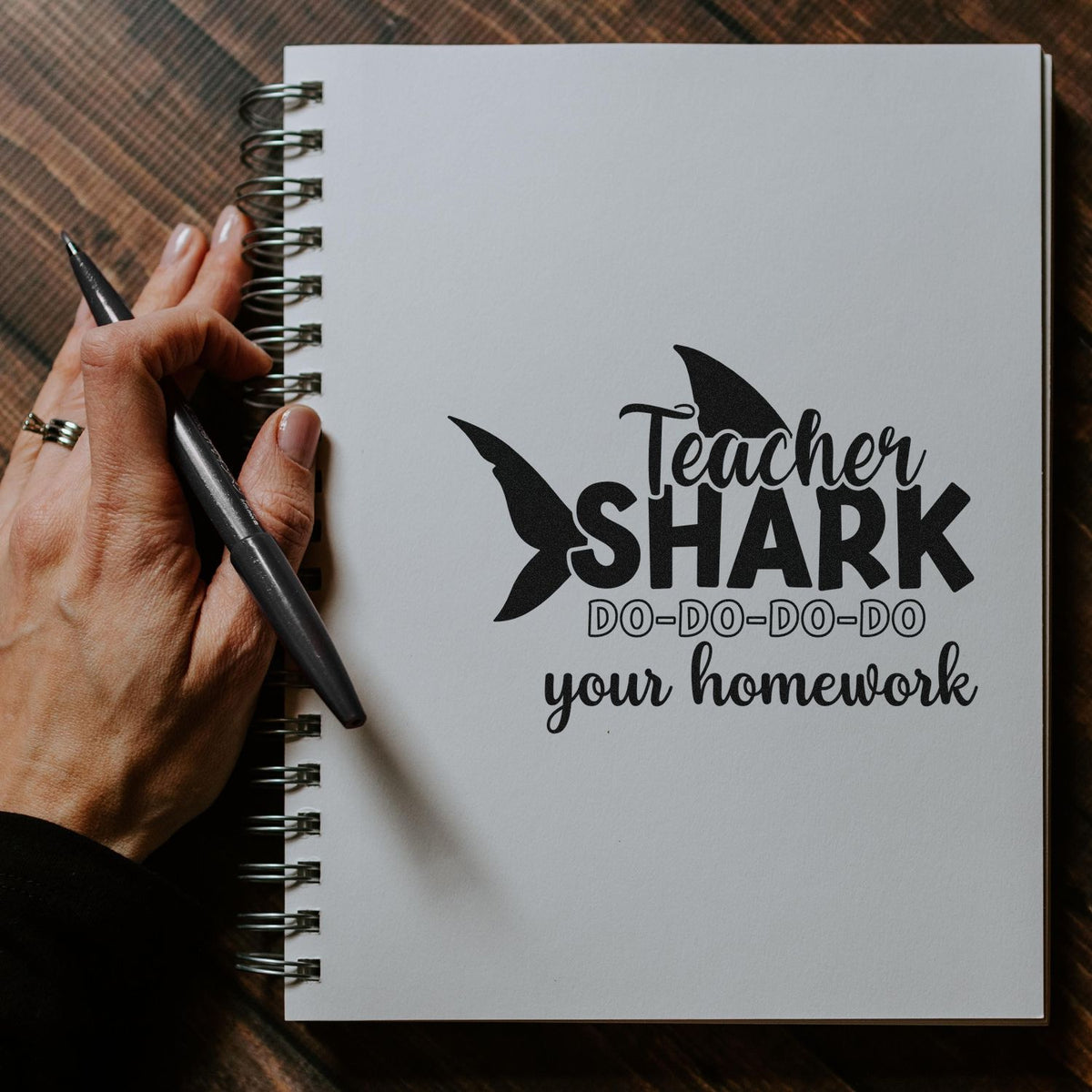 Black teacher shark stamp and seal design on a white spiral notebook, held by a hand with a black pen on brown plaid.