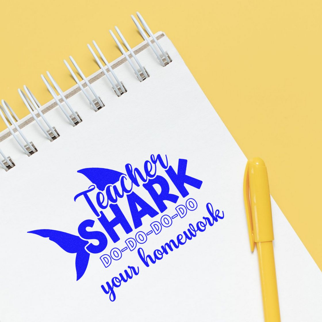 Blue teacher shark stamp and seal design on white notebook with yellow pen on yellow background.