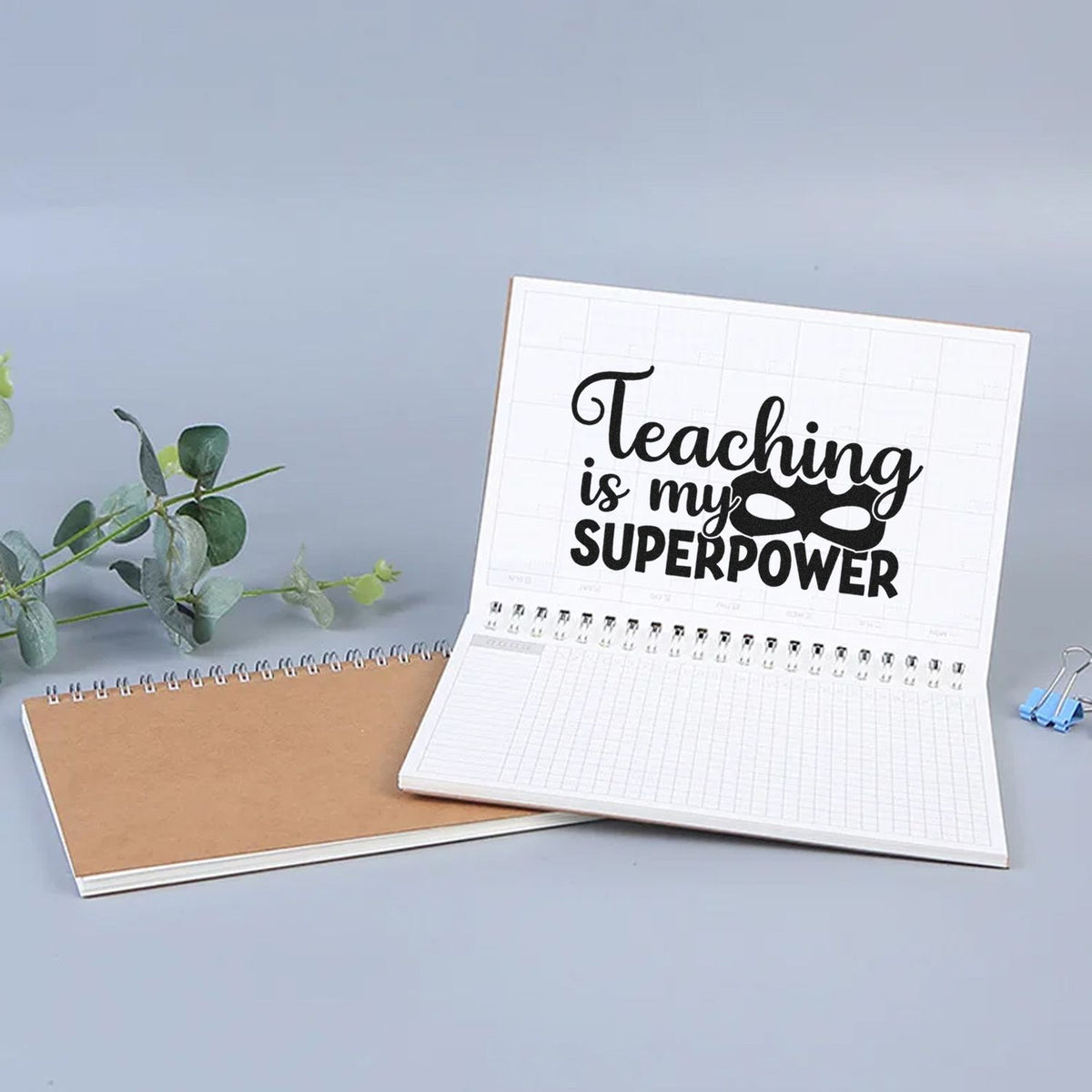 Black teacher stamp reading Teaching is my superpower on white planner pages, with tan notebook and gray backdrop.