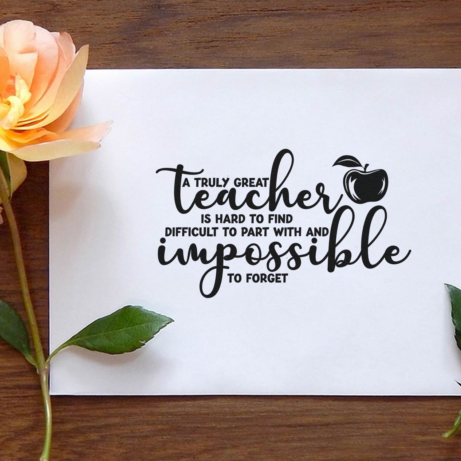 Black teacher stamp and seal on white paper, with apple icon, on a wood table beside a peach flower and green leaves.