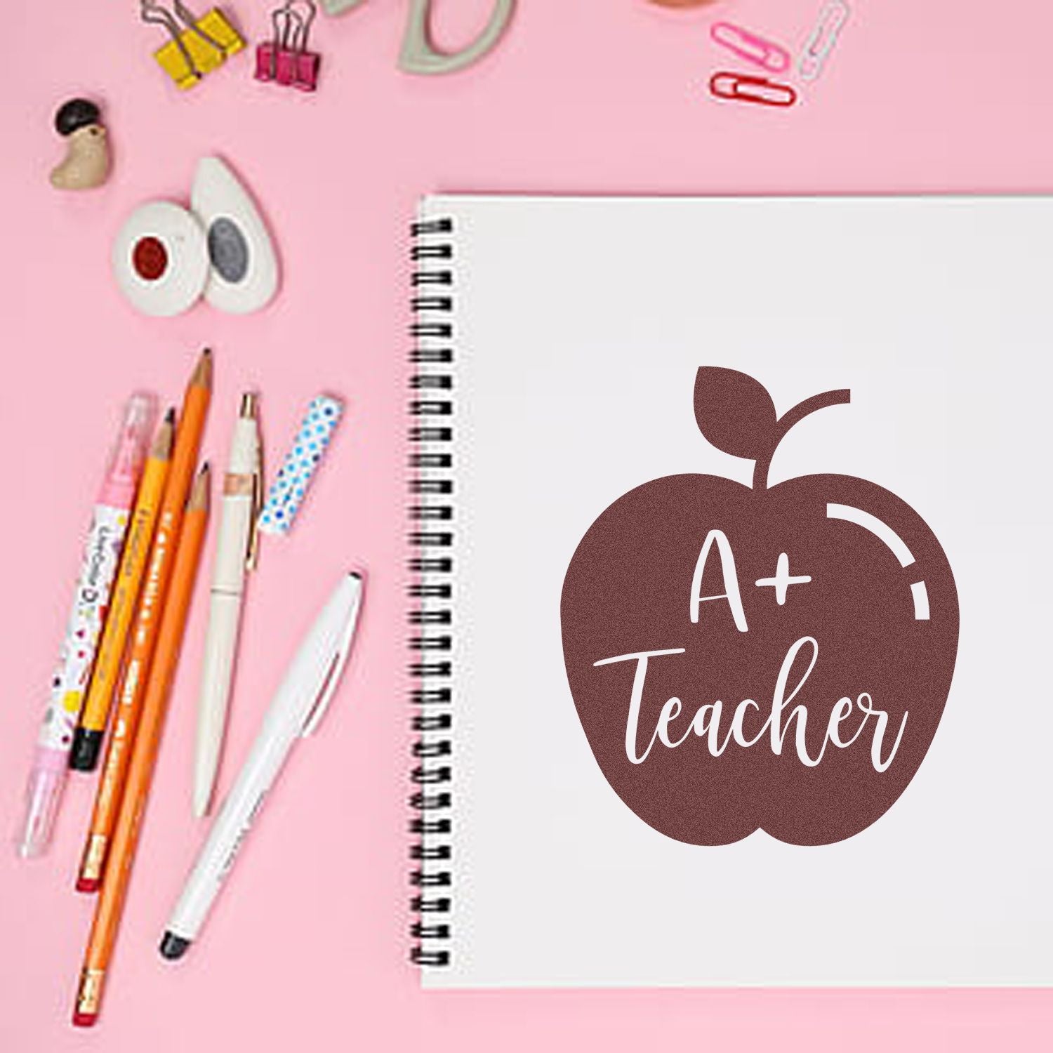 Brown apple-shaped A+ Teacher stamp on white spiral notebook with pink background and colorful stationery.