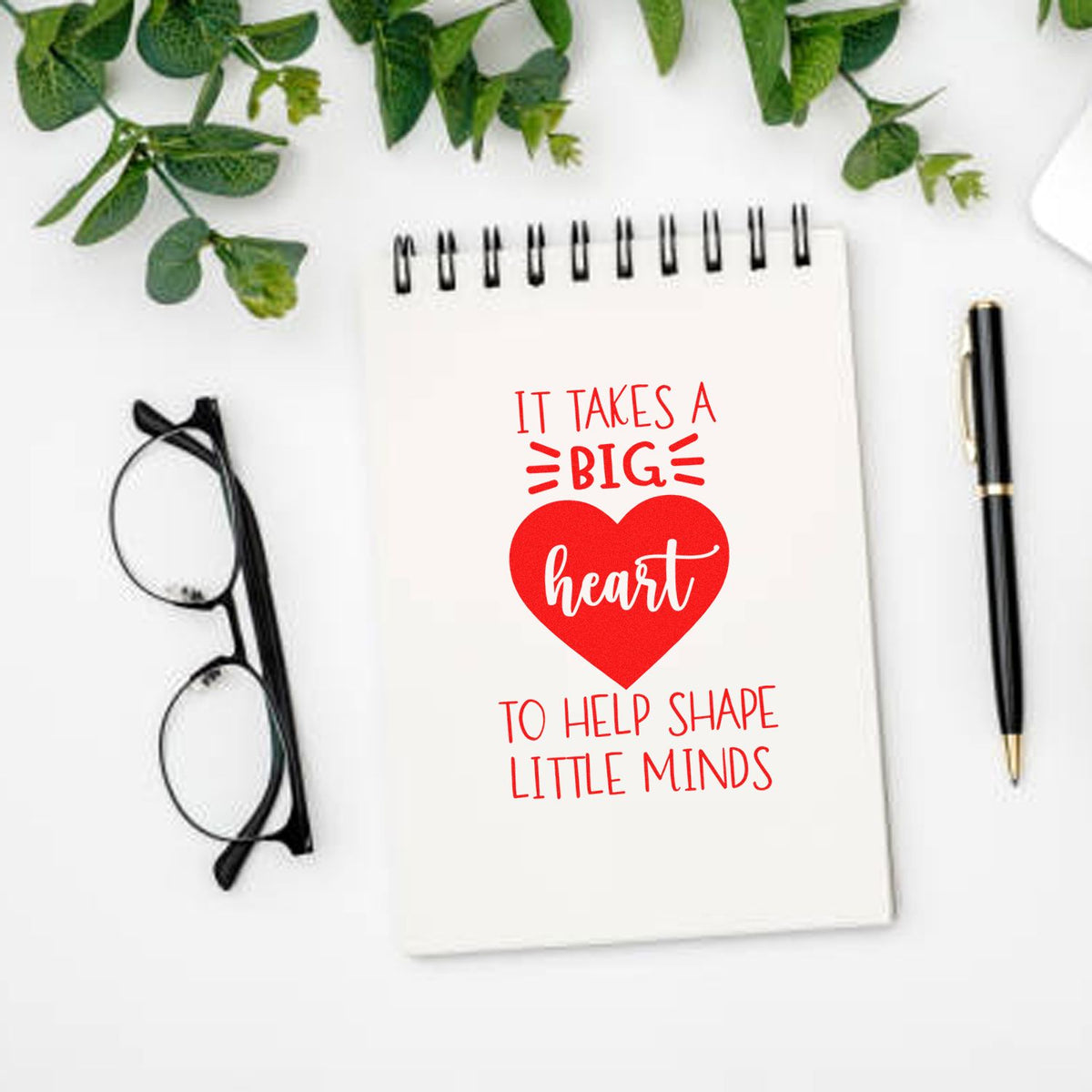 Teacher appreciation rubber stamp with red heart and text on a white spiral notebook, beside black glasses and pen.