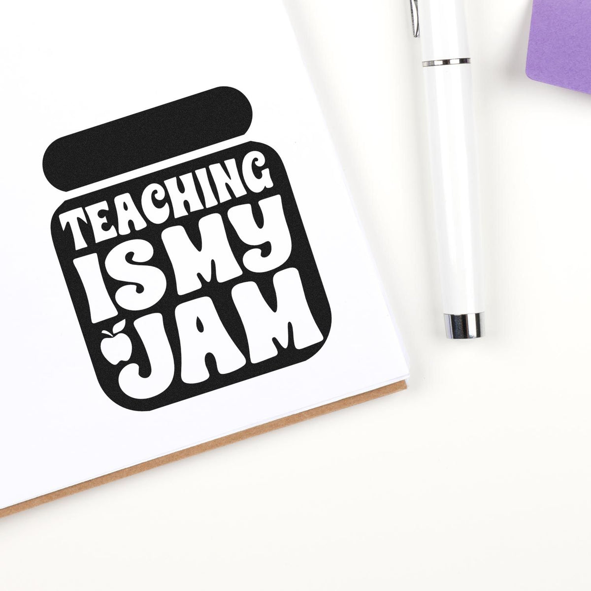 Black teaching stamp seal with Teaching Is My Jam text on white paper, beside a white pen and purple note.