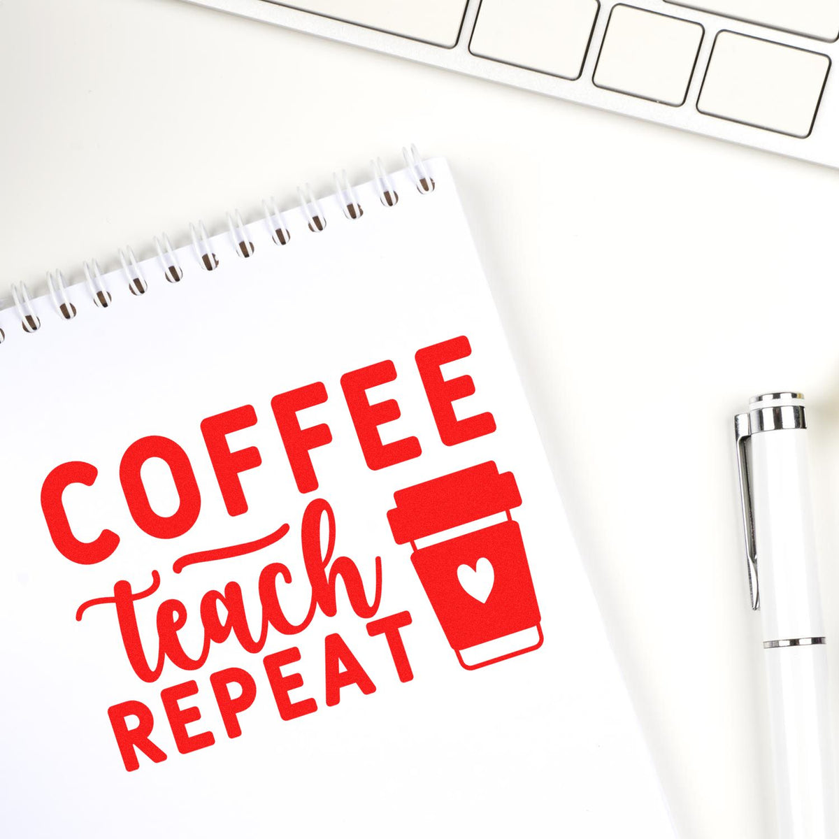 Red custom stamp imprint reading Coffee Teach Repeat with takeaway cup icon on white notebook, beside silver pen and keyboard