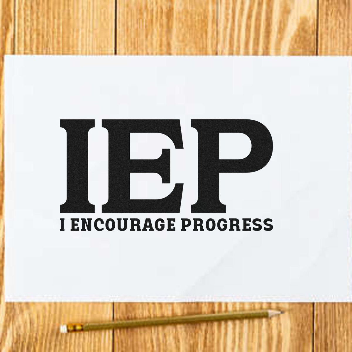 Black IEP rubber stamp reading I ENCOURAGE PROGRESS on white paper, set on a wood surface with a pencil.