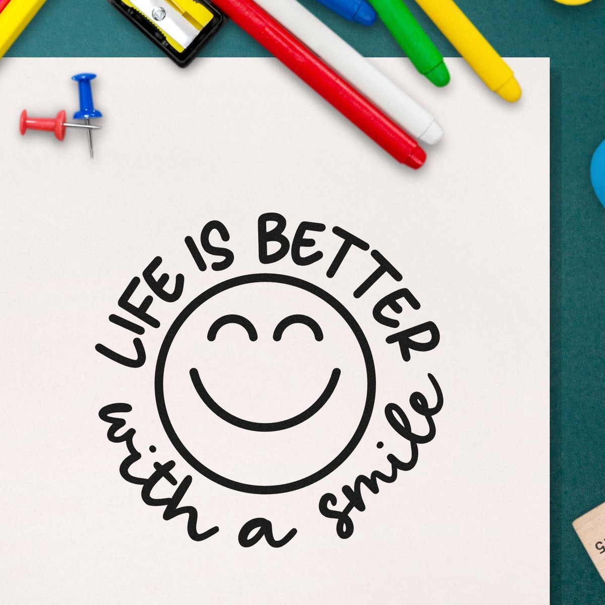 Smiley life-is-better-with-a-smile stamp and seal design on white paper, with colorful pens and push pins.