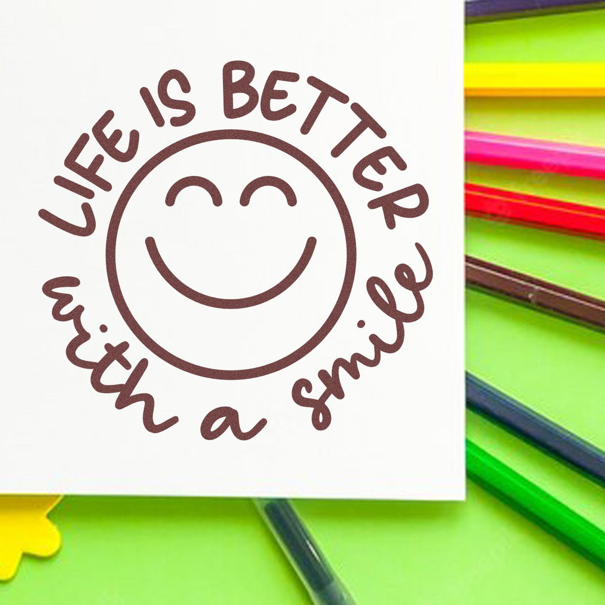 Brown circular stamp seal with smiley face and Life is better with a smile on white, over neon pencils.