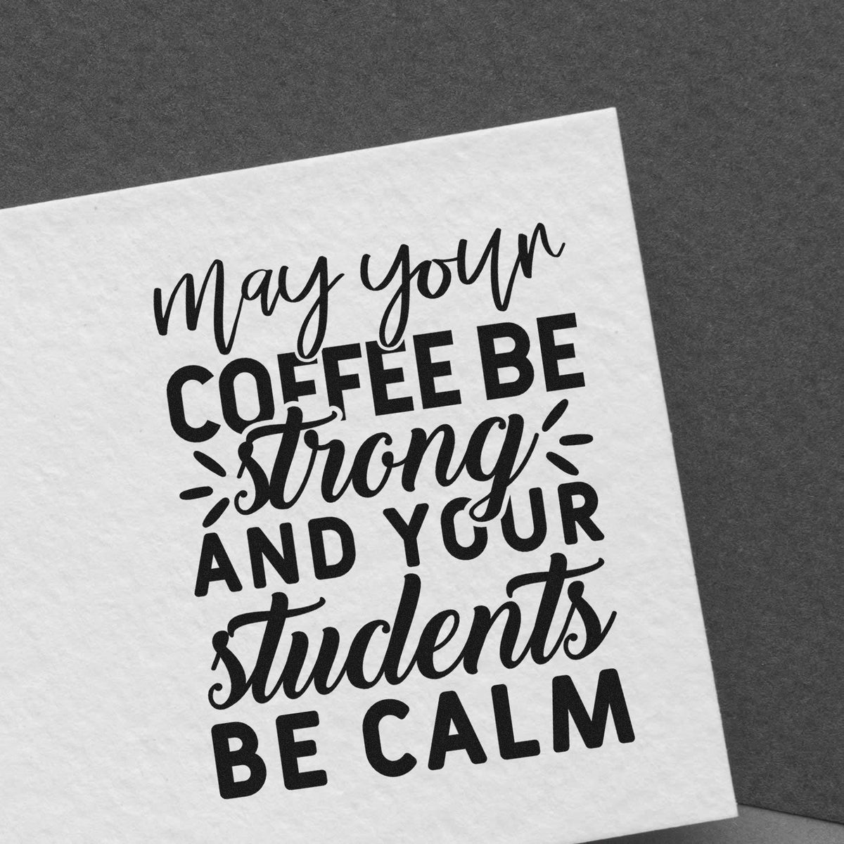 Black stamp-style quote on white paper: May your coffee be strong and your students be calm on a dark gray background.