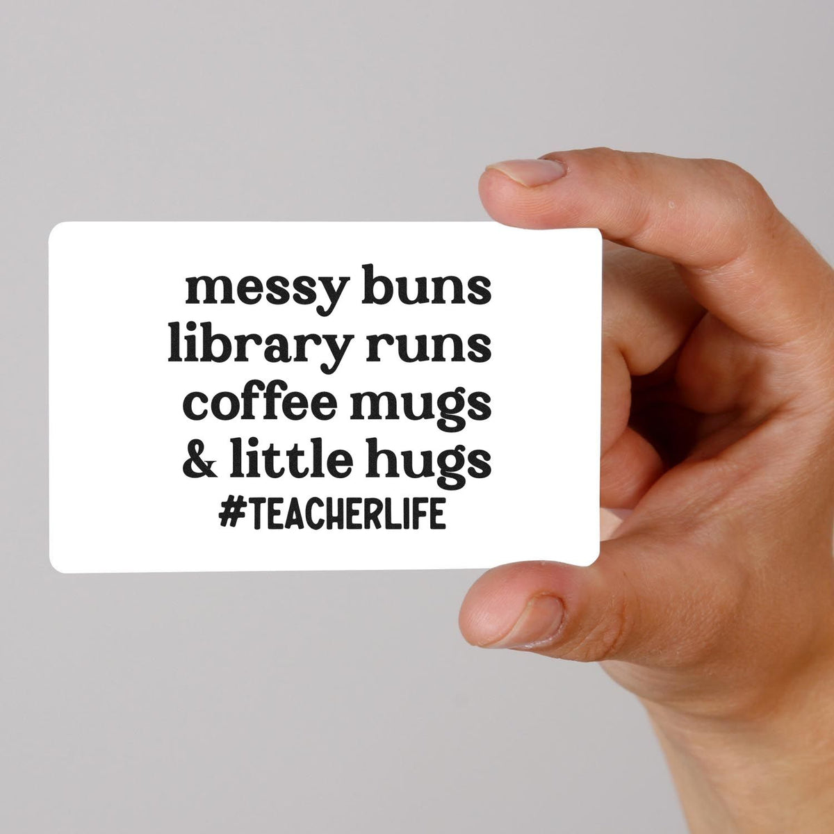 Hand holding a white custom stamp/seal card with black teacher-life text: messy buns, library runs, coffee mugs & little hugs.