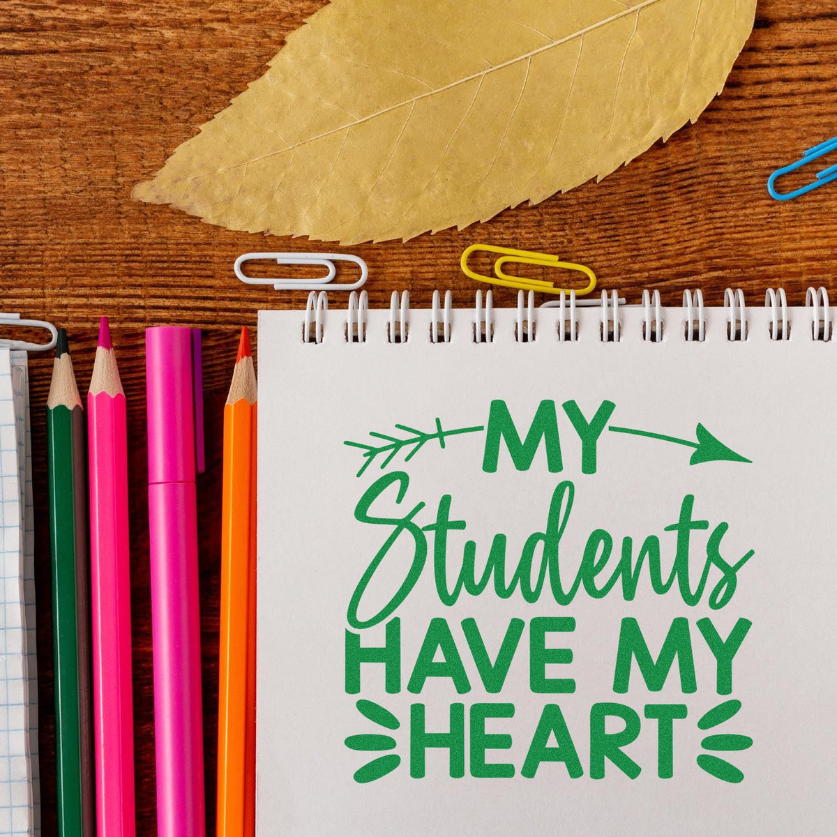 Green stamp seal on a white notebook reading My Students Have My Heart, with colorful pencils, paper clips, and a leaf on wood.