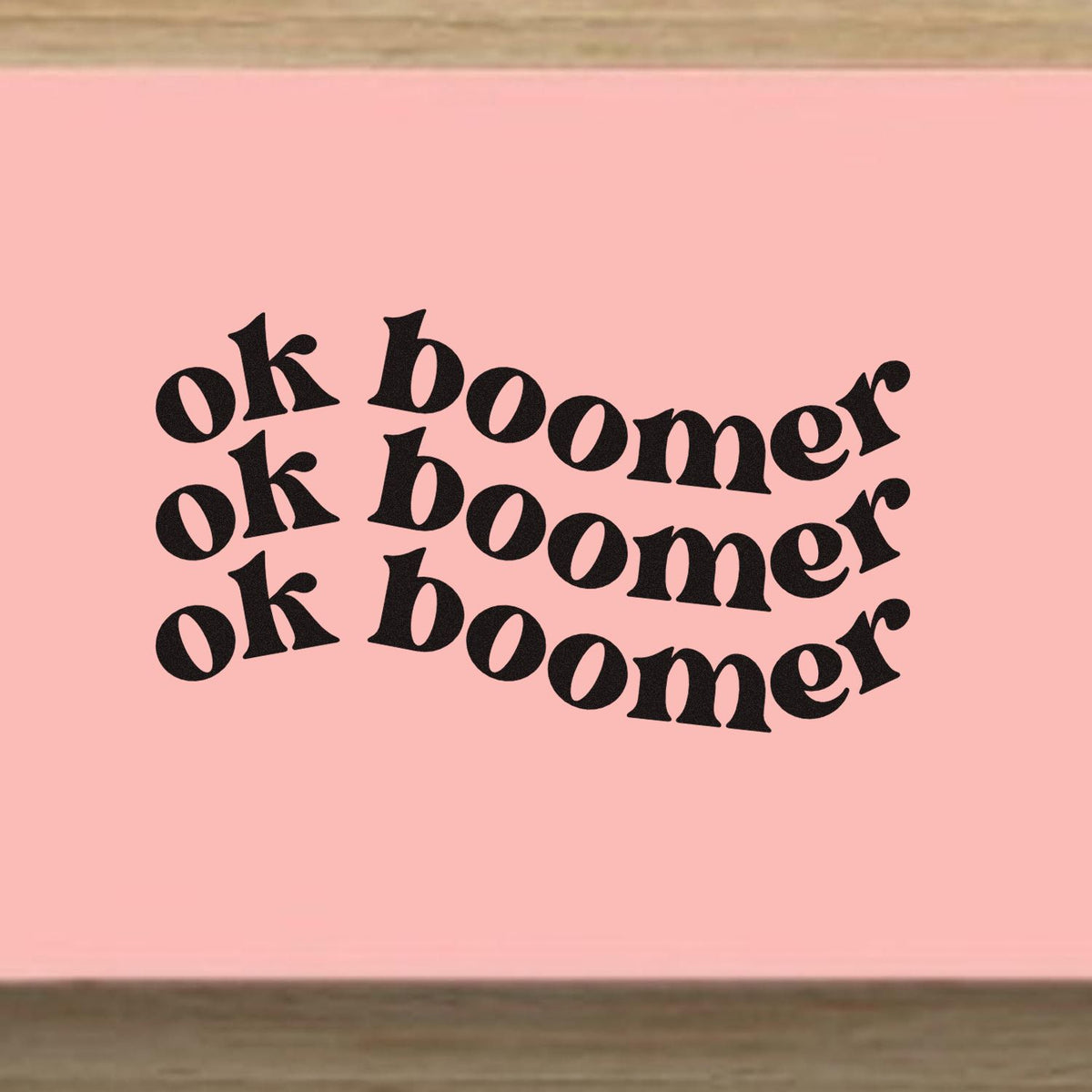 Pink rubber stamp seal with three black ok boomer impressions in bold retro lettering on a blush pink background.