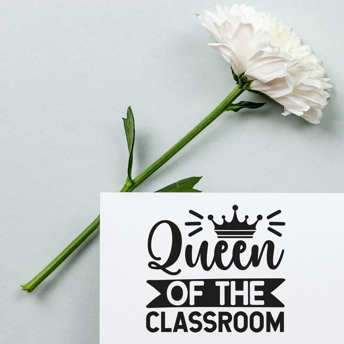White flower beside custom stamp seal reading Queen of the Classroom on a white card.
