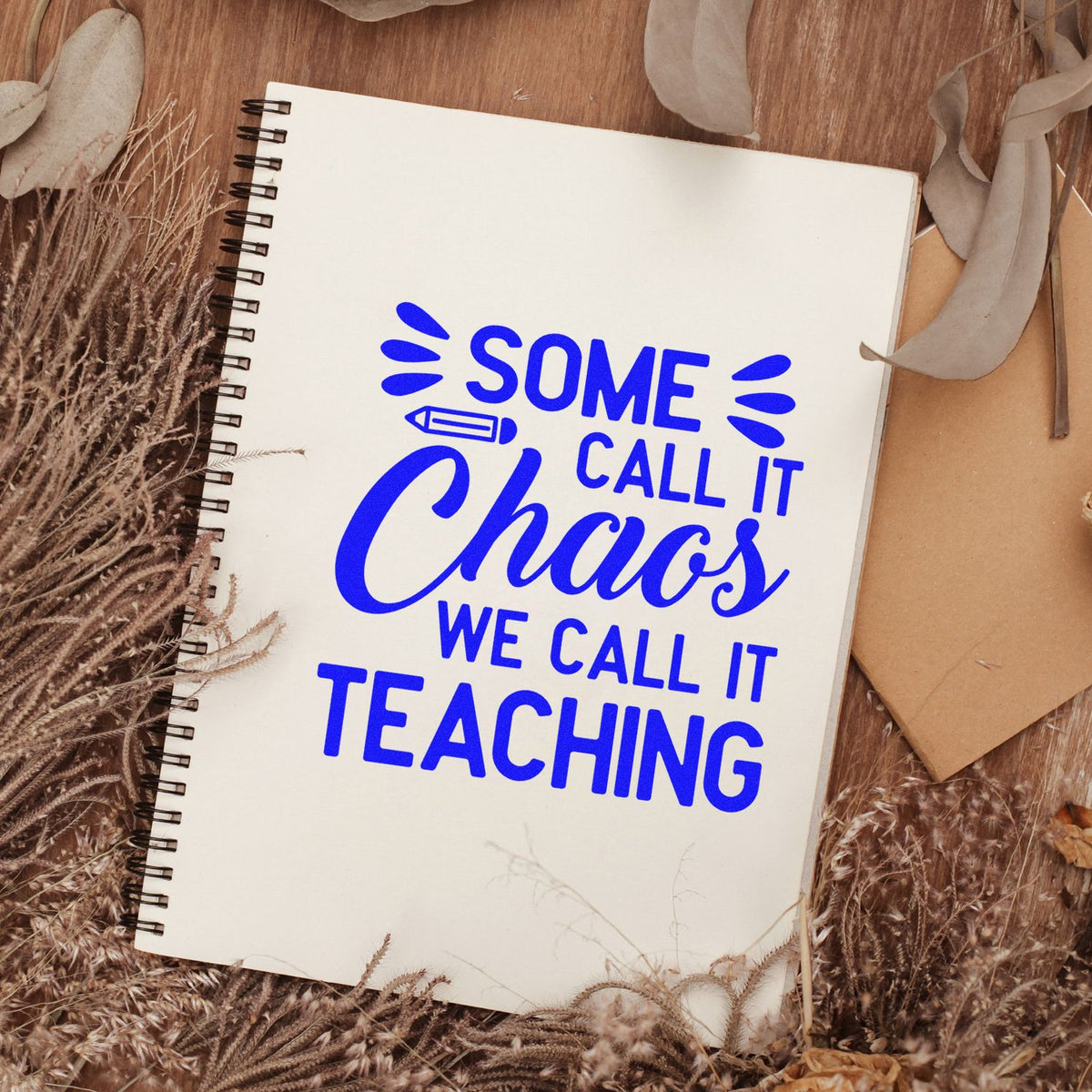 Blue stamp/seal on cream spiral notebook reading Some call it chaos, we call it teaching, with dried botanicals.