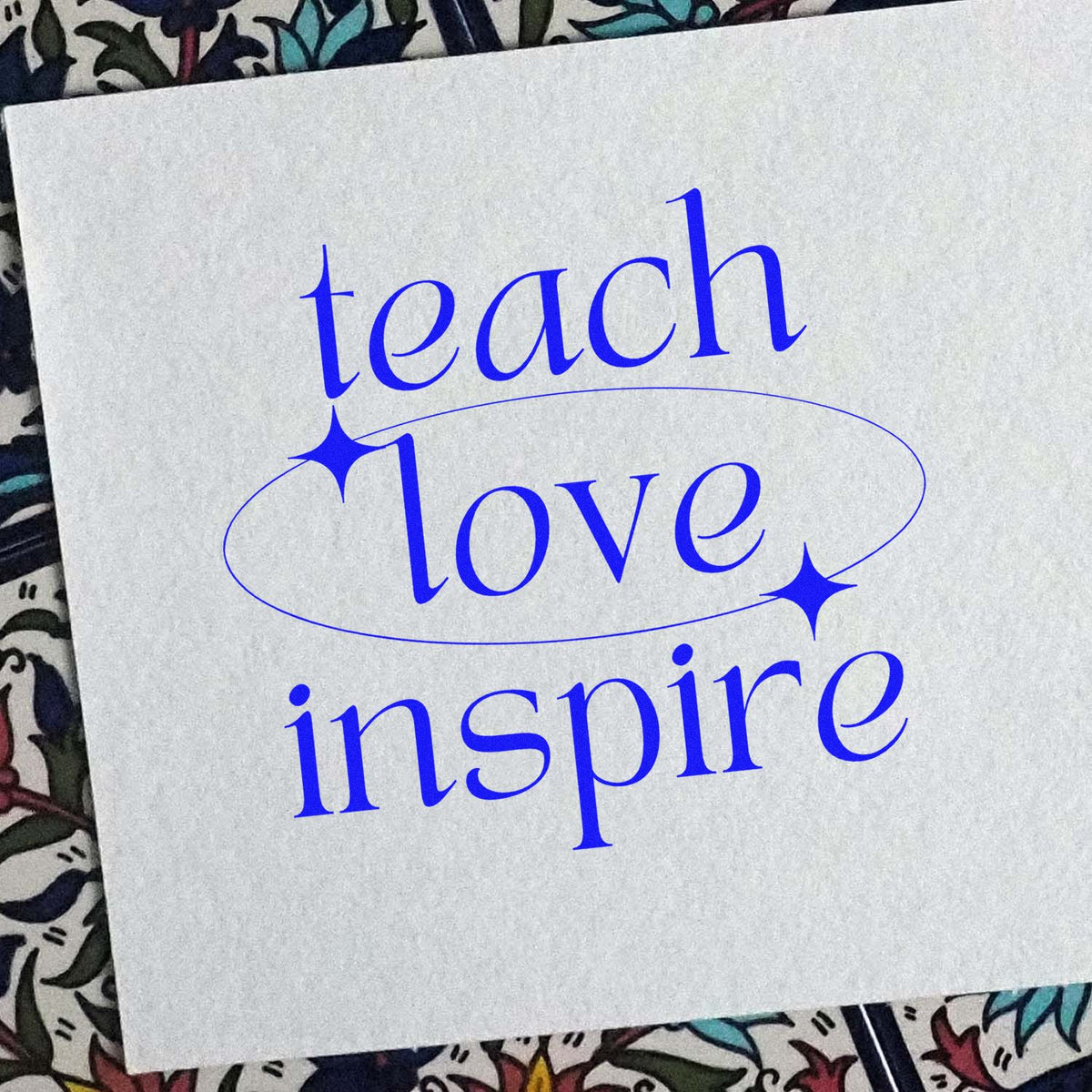 Blue rubber stamp design reading teach love inspire with oval seal and sparkle accents on white paper.