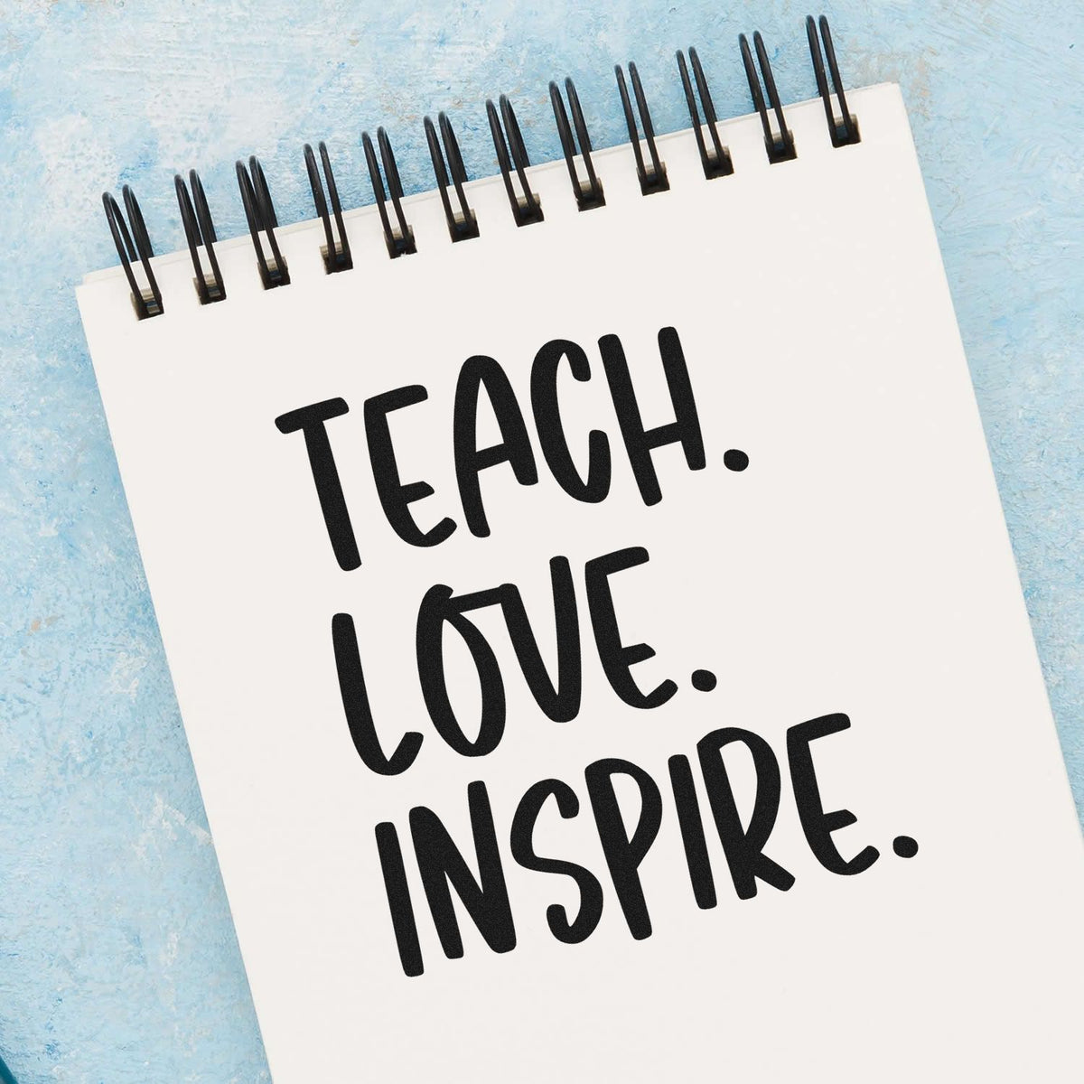 Teacher stamp and seal notepad with black Teach. Love. Inspire. text on a white spiral pad and blue textured background