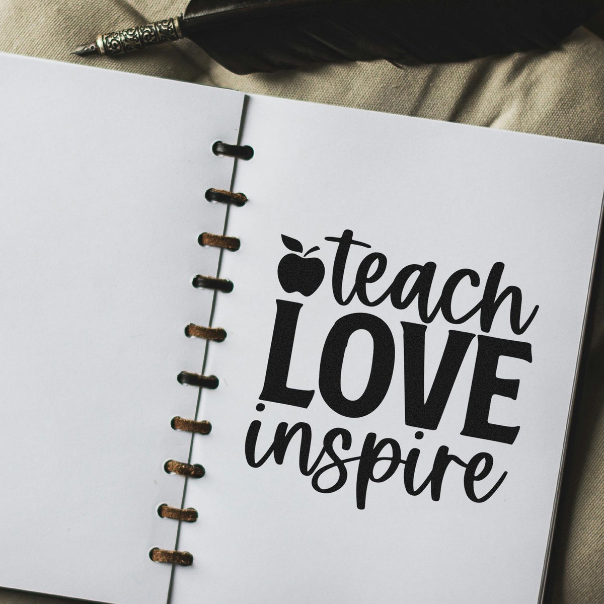 Black stamp-style teach love inspire on white spiral-bound notebook, with seal-like script and quill pen.