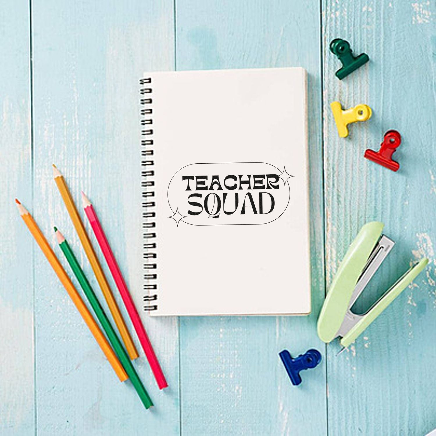 Spiral notebook with black Teacher Squad stamp, colored pencils, binder clips, and pastel stamp/seal tools on light blue wood.
