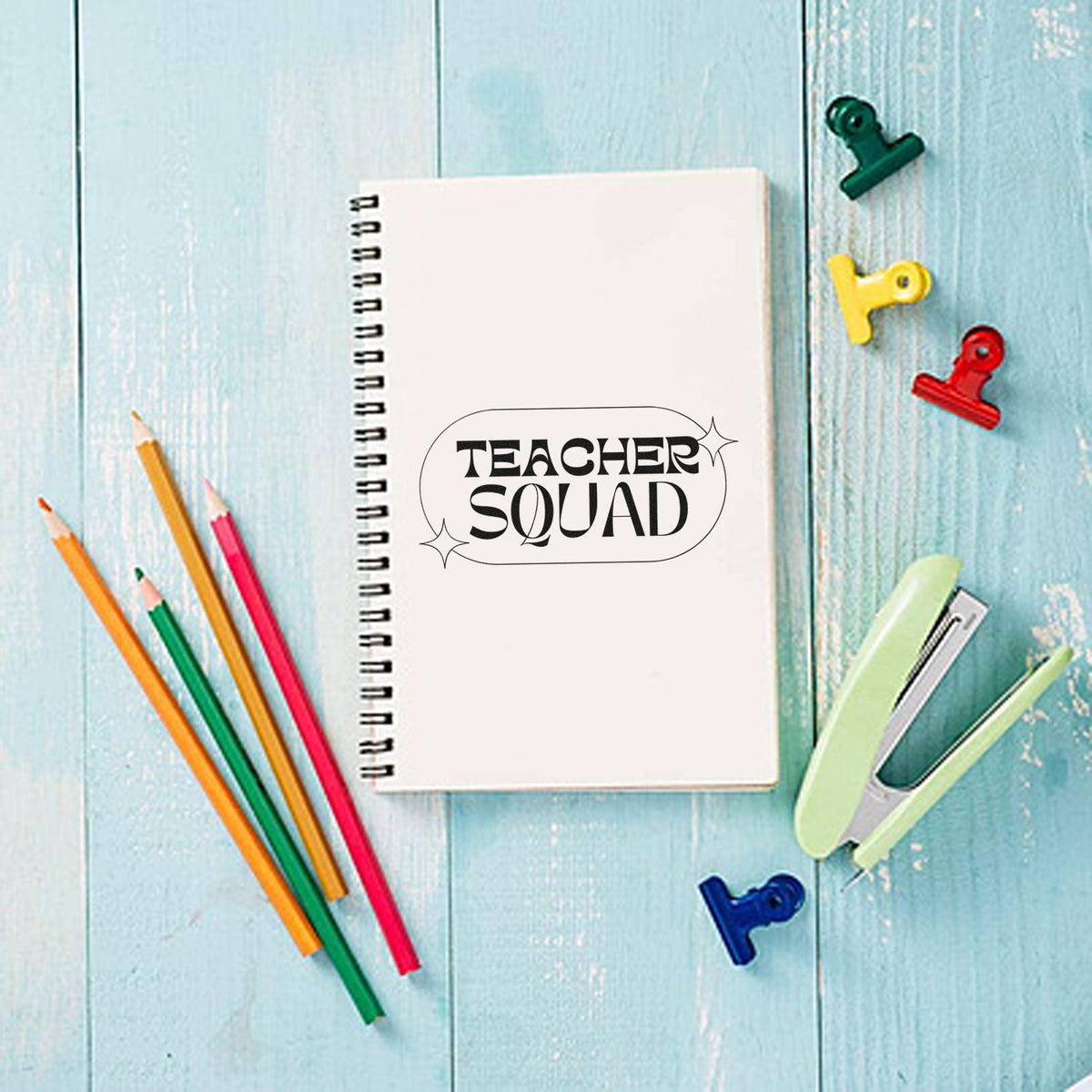 White spiral notebook featuring a black Teacher Squad stamp seal, with colorful pencils, binder clips, and stapler on blue wood.