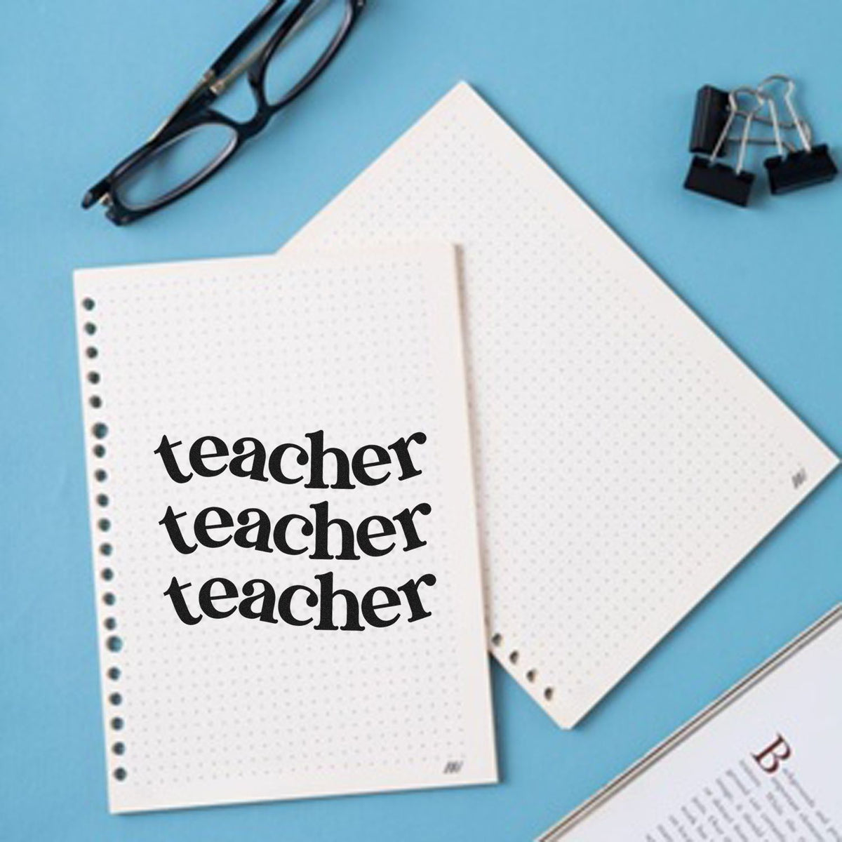 Teacher stamp and seal on white dotted notebooks, with black glasses and binder clips on a blue background.