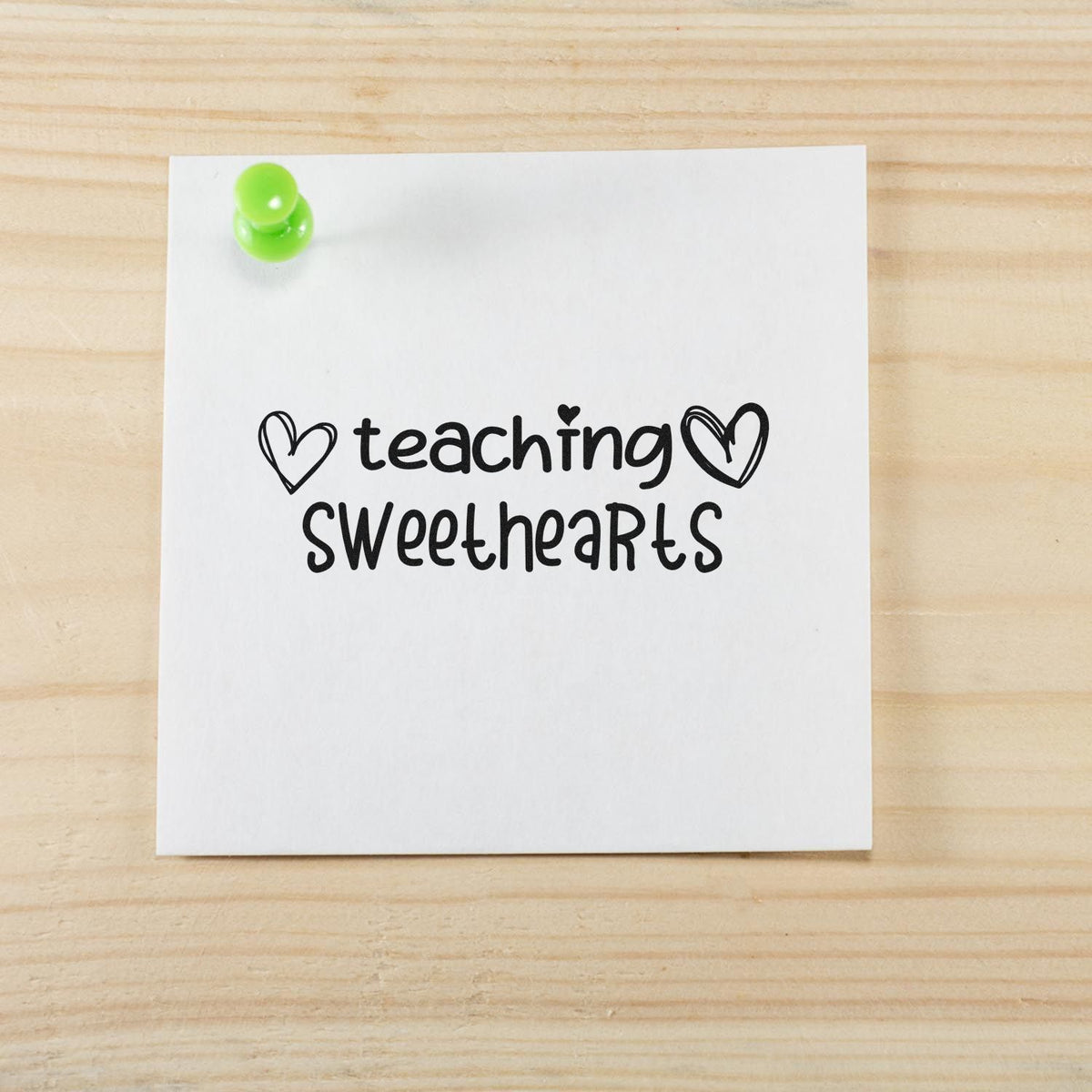 Black teaching sweethearts rubber stamp with hearts on white paper, pinned with bright green pushpin on light wood.