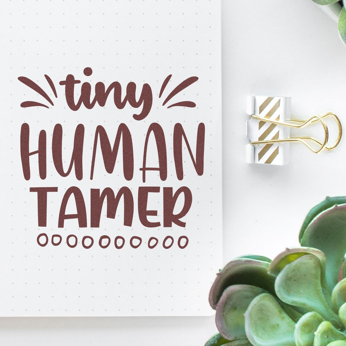 Maroon Tiny Human Tamer stamp and gold-striped seal on dotted notebook page, with green succulent.