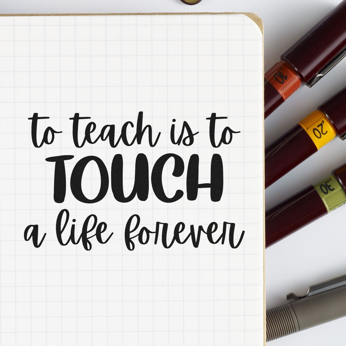 Grid notebook with black hand-lettered quote beside burgundy stamp and seal markers with orange, yellow, green, and gray bands.