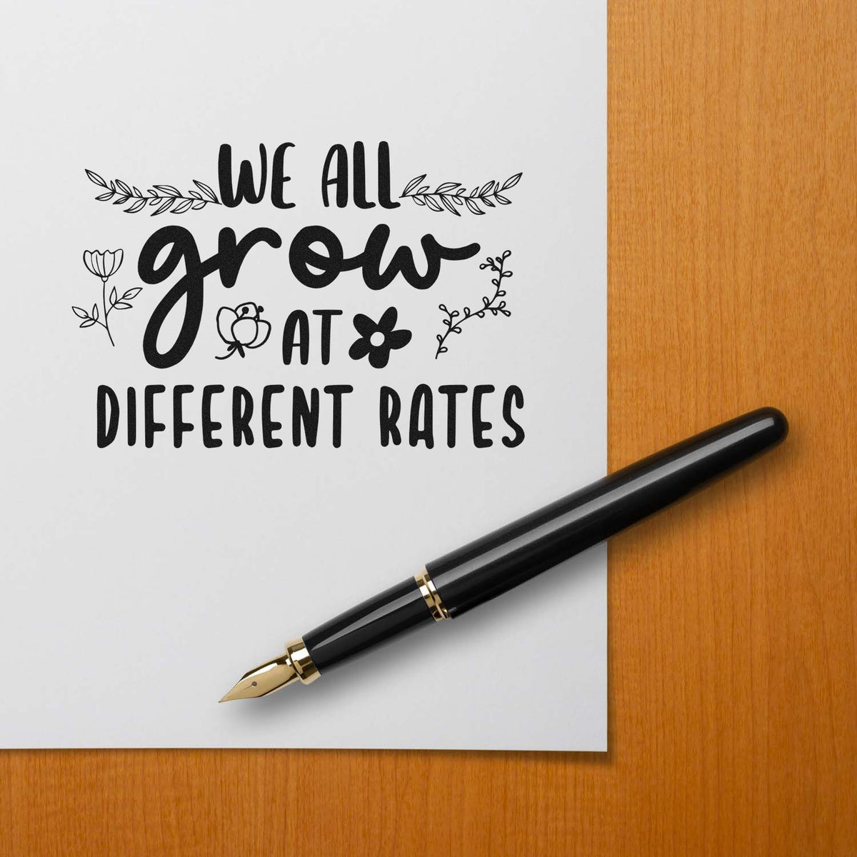 Custom stamp and seal design with black floral quote We all grow at different rates on white paper, beside a black fountain pen.