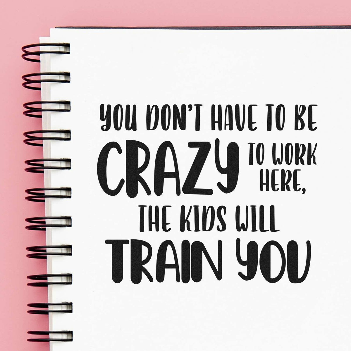 Black stamp-style quote on a white spiral notebook with pink background: You dont have to be crazy to work here, the kids will train you.