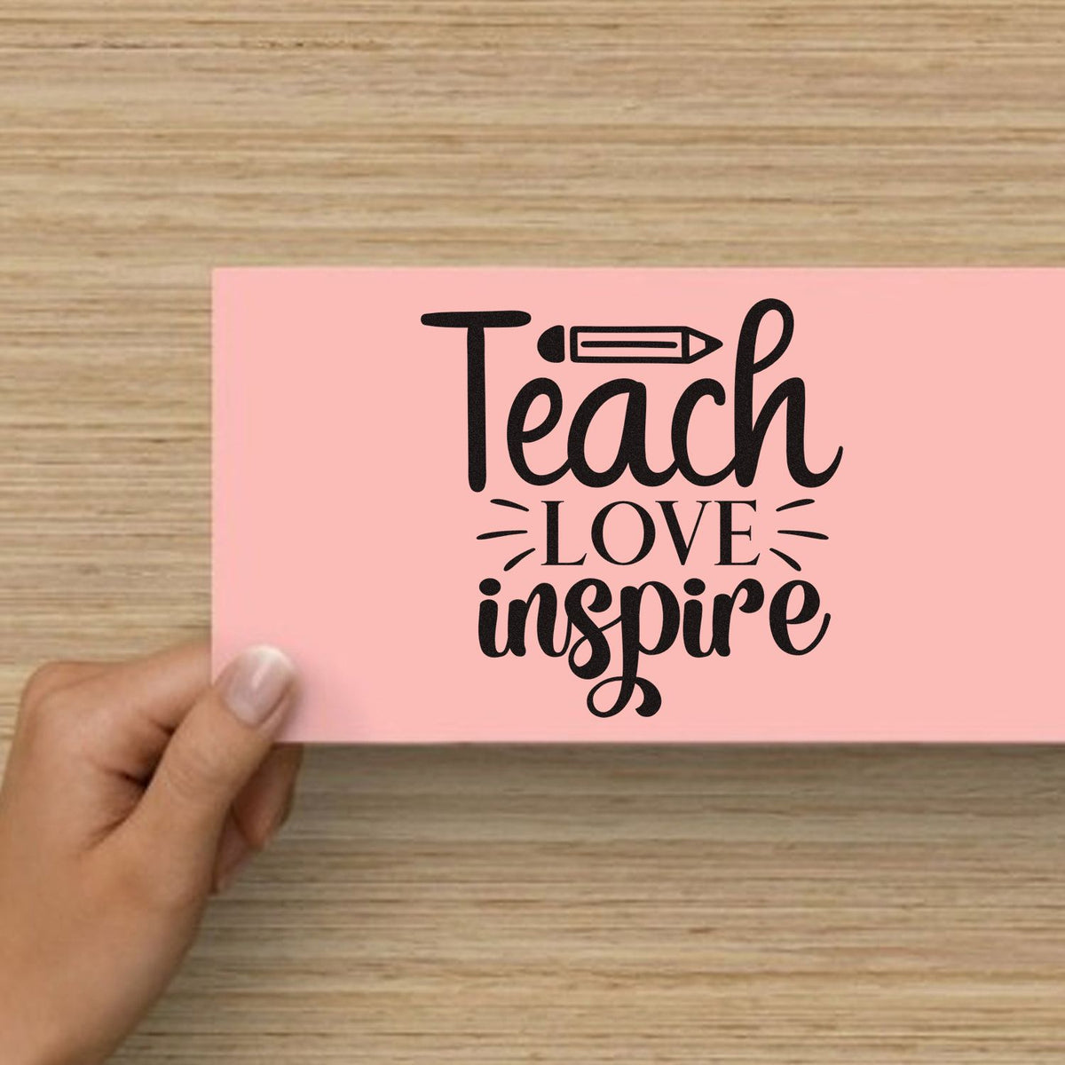Pink custom stamp seal card with black Teach Love Inspire lettering, pencil icon, and hand holding it on wood background.
