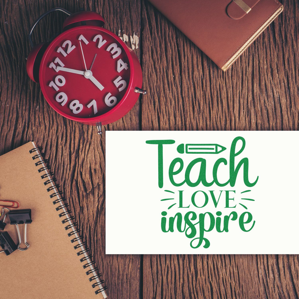 White stamp seal card with green Teach Love Inspire lettering on wood desk, beside a red alarm clock and brown notebook.