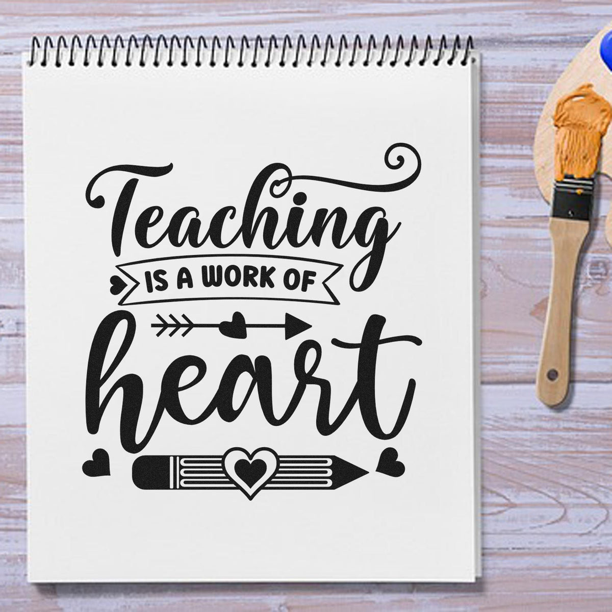 Black teaching heart stamp on white spiral notebook, with pencil and hearts; wooden brush seal product at right.
