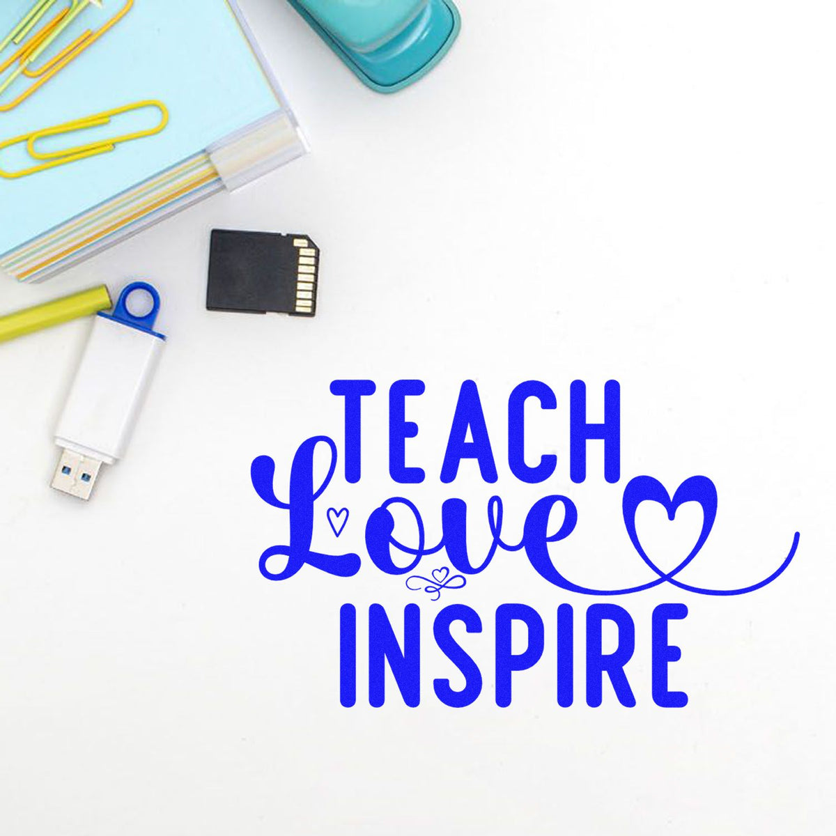 Blue custom stamp and seal with Teach Love Inspire and heart, beside paper clips, USB drive, and SD card.
