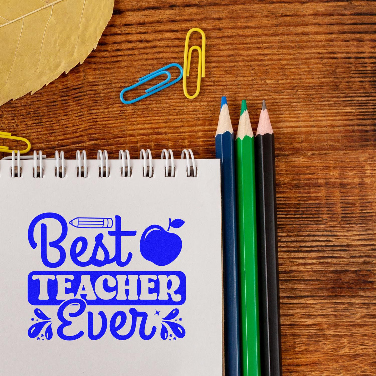 Blue Best Teacher Ever teacher stamp design on spiral notebook, with colored pencils, paperclips, and wood desk.