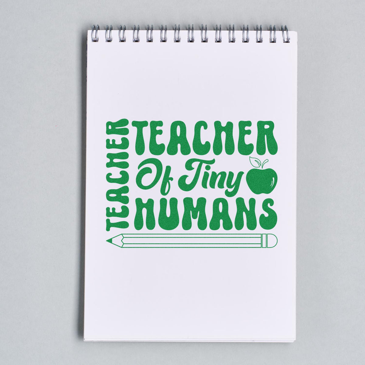 Green teacher stamp seal reading Teacher of Tiny Humans with apple and pencil on white notepad.