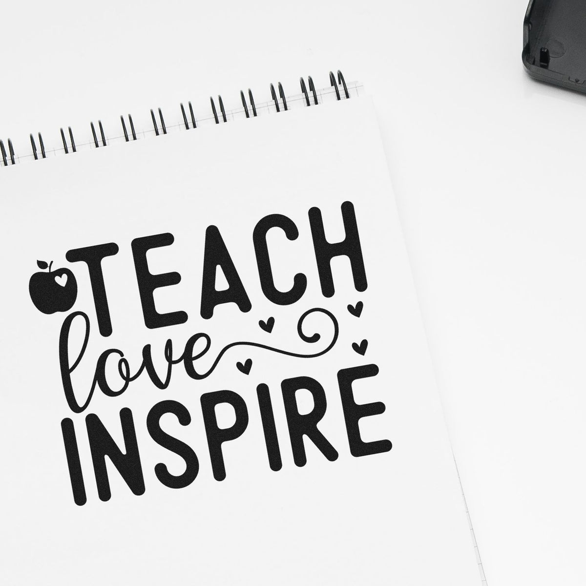 Black teacher stamp/seal design on white spiral notebook, with apple, hearts, and Teach love inspire text.