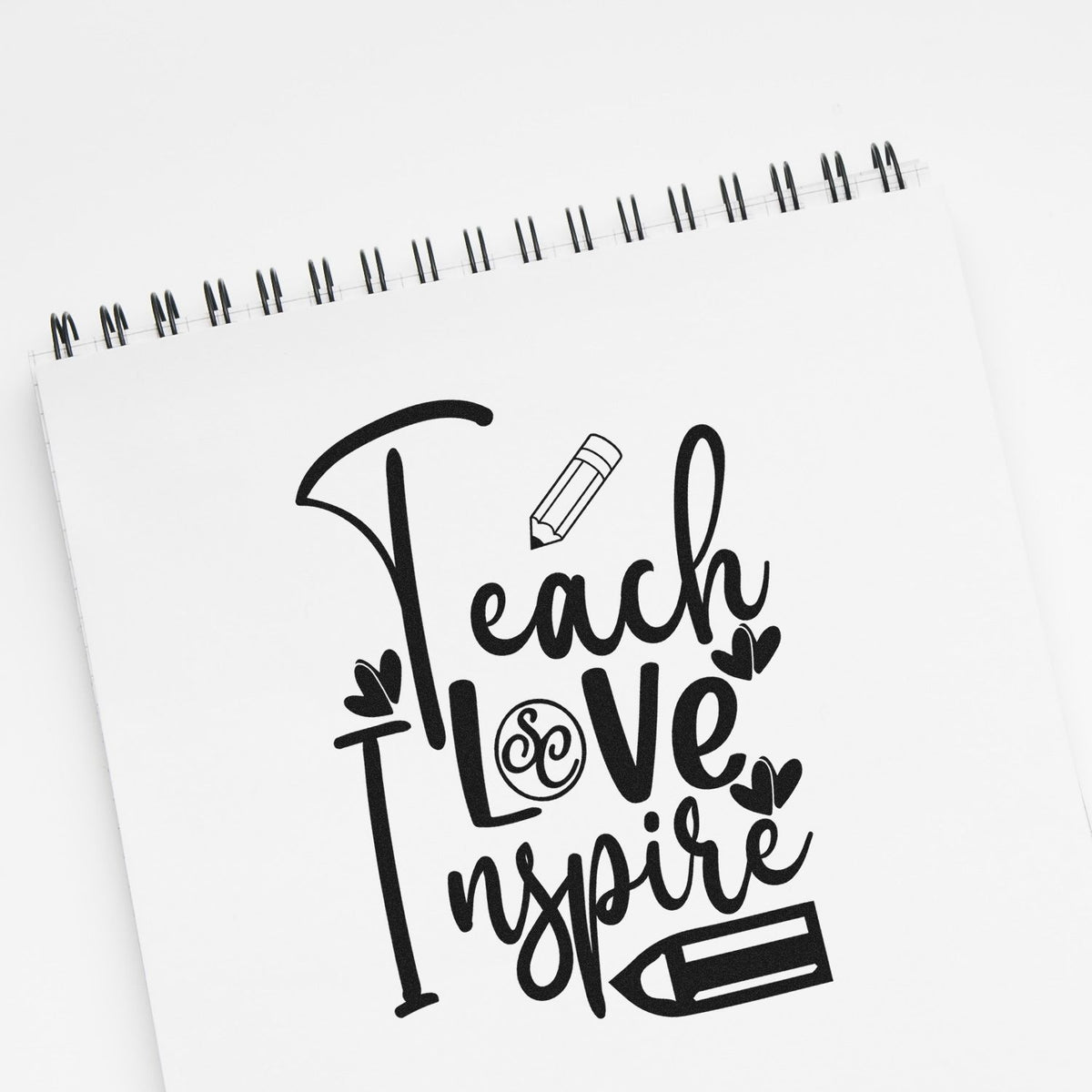 Black Teach Love Inspire stamp and seal design on white spiral notebook with pencil and hearts.
